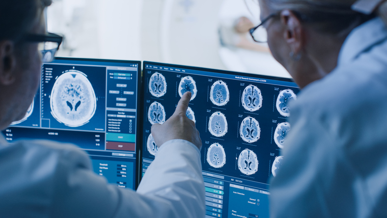 In Control Room Doctor And Radiologist Discuss Diagnosis While Watching Procedure And Monitors Showing Brain Scans Results, In The Background Patient Undergoes Mri Or Ct Scan Procedure.